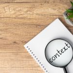 A magnifying glass is positioned over the word "context" written on a notebook, emphasizing the importance of understanding context.