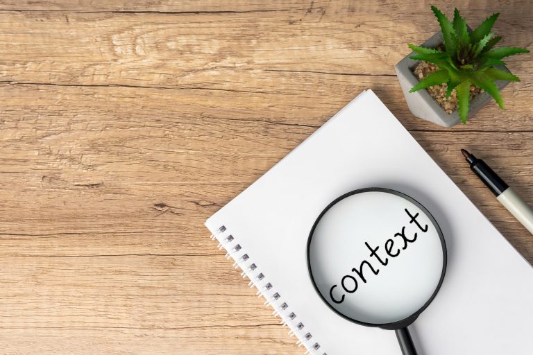 A magnifying glass is positioned over the word "context" written on a notebook, emphasizing the importance of understanding context.